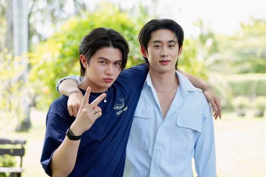 Foto dos atores tailandeses William Jakrapatr Kaewpanpong e Est Supha Sangaworawong abraçados e sorrindo ao ar livre em um parque. William usa uma camiseta azul e faz o sinal de paz.