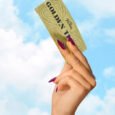 Mão erguendo o Bilhete Dourado de Willy Wonka contra um céu azul com nuvens, em referência ao reality show Wonka: O Bilhete Dourado da Netflix.